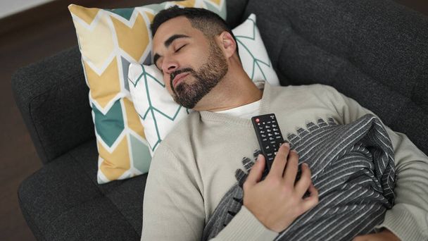 Young hispanic man holding tv remote control lying on sofa sleeping at home - Photo, Image