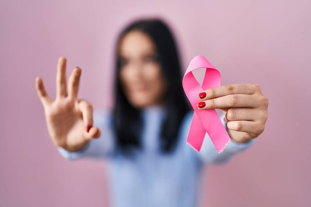Іспаномовна жінка, яка тримає рожеву ракову стрічку, малює пальцями, усміхається, жестикулює чудовий символ.  - Фото, зображення
