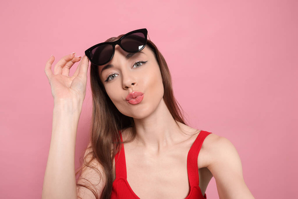 Beautiful young woman with sunglasses blowing kiss while taking selfie on pink background - Fotoğraf, Görsel