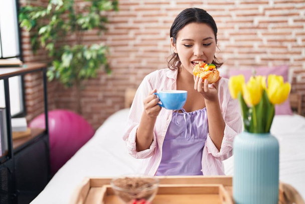 Young hispanic woman having gift breakfast sitting on bed at bedroom - Foto, Imagem