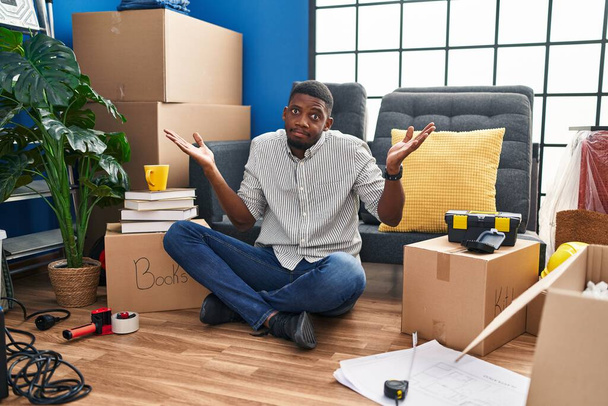 Afroamerikaner sitzt ratlos und verwirrt mit erhobenen Armen und Händen auf dem Boden in seinem neuen Zuhause. Zweifelhaftes Konzept.  - Foto, Bild