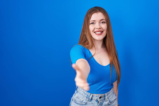 Femme rousse debout sur fond bleu souriant amical offrant poignée de main comme salutation et accueil. entreprise prospère.  - Photo, image