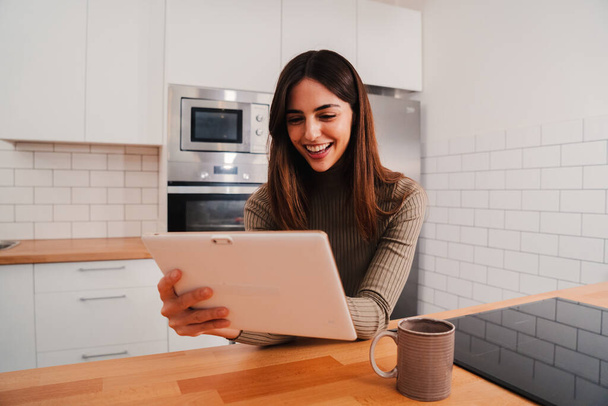 Mutfakta oturan kablosuz konforu kullanarak internette gezinebileceği bir tablet cihazı olan mutlu bir beyaz kadın. Gülümseyen ve online alışveriş yapmaktan zevk alan genç bir kadını izole et. Yüksek kalite - Fotoğraf, Görsel
