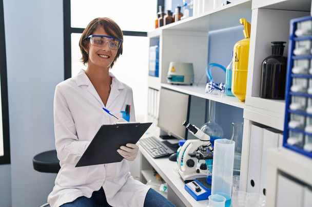 Young beautiful hispanic woman scientist smiling confident writing report working at laboratory - Foto, afbeelding