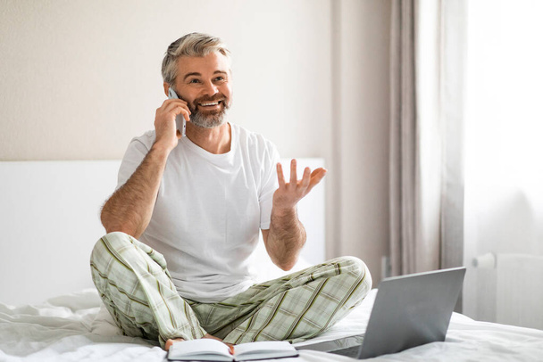 Happy handsome grey-haired bearded mature businessman wearing pajamas sitting on bed, using computer laptop, talking on phone, holding pencil, looking at copy space and smiling - Fotoğraf, Görsel