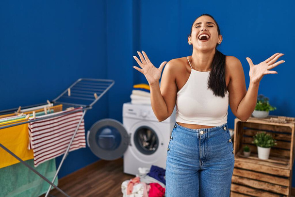 Mujer hispana joven en la lavandería celebrando loco y loco por el éxito con los brazos levantados y los ojos cerrados gritando emocionado. concepto ganador  - Foto, imagen