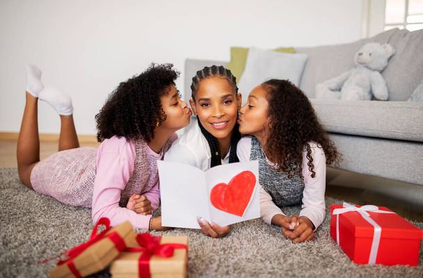 African American Preteen Girls Kissing Mom Congratulating Her On Mothers Day Lying On Floor At Home. Young Mommy Smiling To Camera Receiving Gifts From Kids Celebrating Family Holiday Together - 写真・画像
