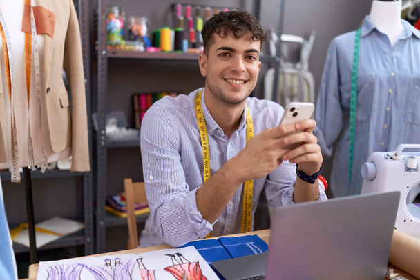 Young hispanic man tailor using smartphone and laptop at atelier - Photo, Image