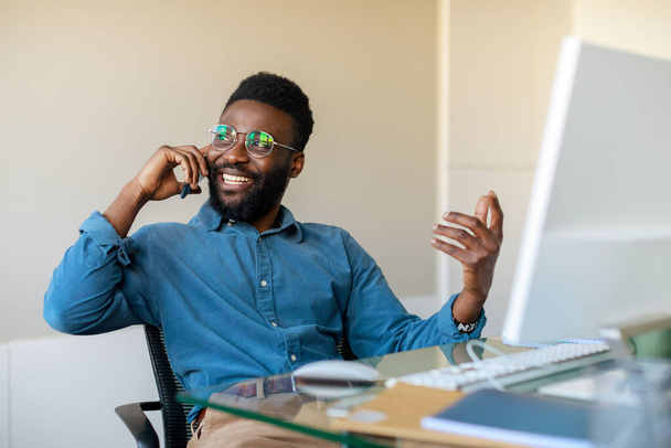 Udany czarny biznesmen rozmawiający przez telefon, prowadzący rozmowę biznesową i uśmiechnięty, siedzący w biurze w miejscu pracy. Pozytywny mężczyzna przedsiębiorca mówi przez telefon komórkowy i gestykulacji - Zdjęcie, obraz