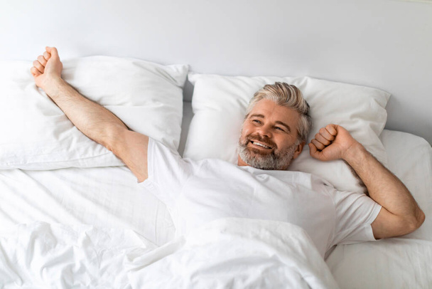 Boldog pozitív jóképű ősz hajú szakállas európai középkorú férfi fehér pólóban nyújtózik az ágyban reggel, nézi fénymásolás és mosoly, felülnézet, otthoni belső - Fotó, kép