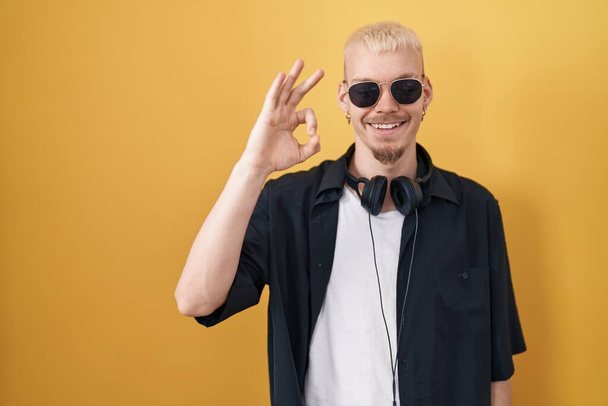 Młody mężczyzna rasy kaukaskiej w okularach przeciwsłonecznych stojący nad żółtym tłem uśmiechający się pozytywnie robiący znak ok ręką i palcami. udana ekspresja.  - Zdjęcie, obraz