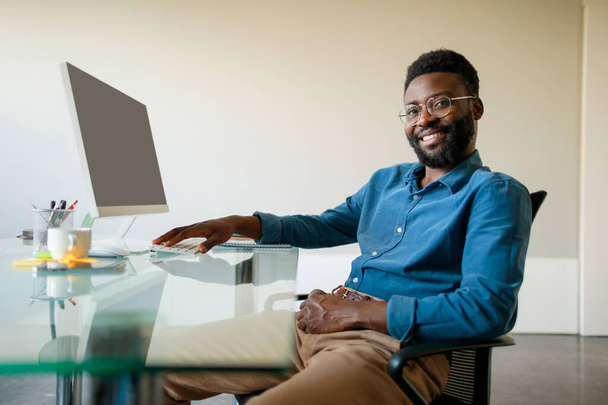Portrait of positive african american male entrepreneur sitting at workdesk in office in front of computer, looking and smiling at camera, free space - Fotó, kép