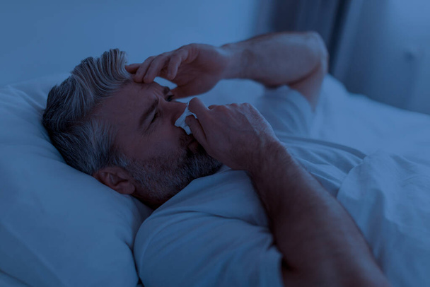 Gros plan d'un homme malade d'âge moyen aux cheveux gris portant un pyjama touchant le nez qui coule avec une serviette, éternuant dans son lit la nuit, touchant la tête, souffrant de rhume, de grippe, de coronavirus - Photo, image