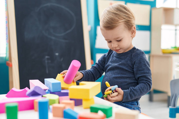 Очаровательный блондин играет с геометрическими блоками, сидя за столом в детском саду - Фото, изображение