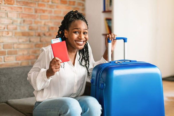 スーツケースの横に座っているアフリカ系アメリカ人の女性を応援し、国際的なパスポートと飛行機のチケットを持ち、カメラで笑顔で旅行の準備をします。夏の旅行のコンセプト - 写真・画像