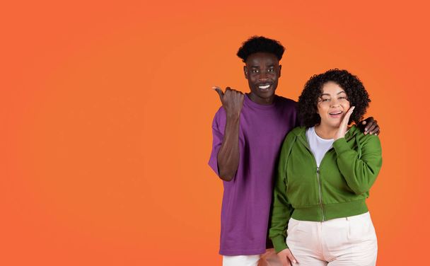 Cute beautiful loving multicultural young couple black man and hispanic woman embracing and showing free space for advertisement over red studio background, mixed race lovers recommending nice offer - 写真・画像