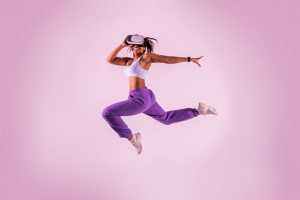 Young black woman working out with 3D technology, jumping mid air while wearing virtual reality headset, pink neon studio background. Sporty lady exploring virtual fitness game. - Foto, Imagem