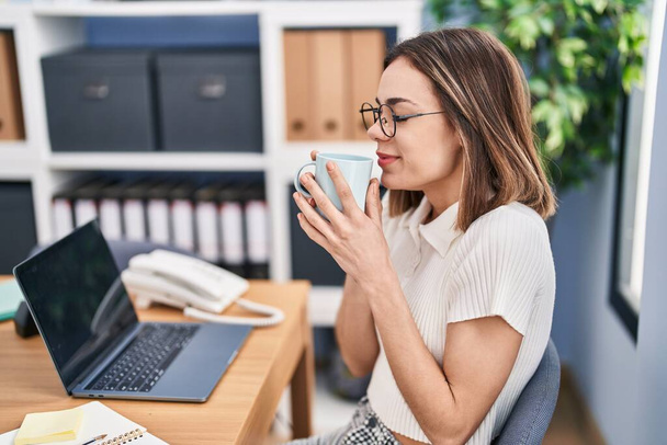 Νεαρή όμορφη ισπανόφωνη επιχειρηματίας χρησιμοποιώντας φορητό υπολογιστή μυρίζοντας καφέ στο γραφείο - Φωτογραφία, εικόνα