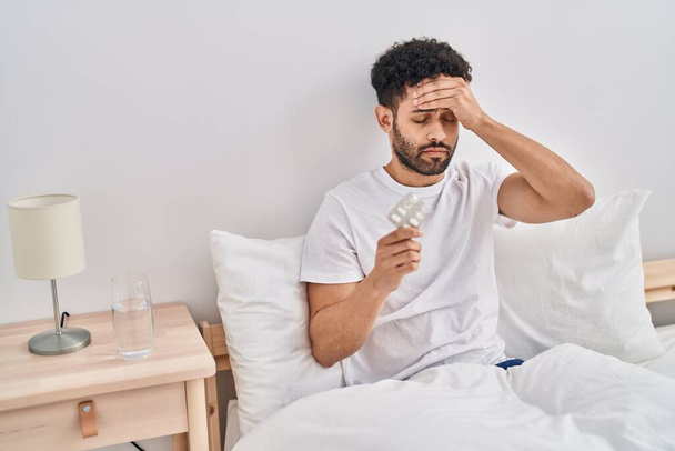Hombre árabe joven tomando pastillas para el dolor de cabeza sentado en la cama en el dormitorio - Foto, imagen