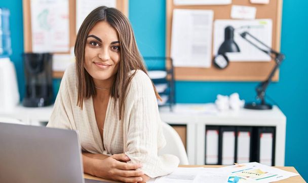 Nuori kaunis latino nainen liiketoiminnan työntekijä hymyilee luottavainen istuu pöydällä toimistossa - Valokuva, kuva