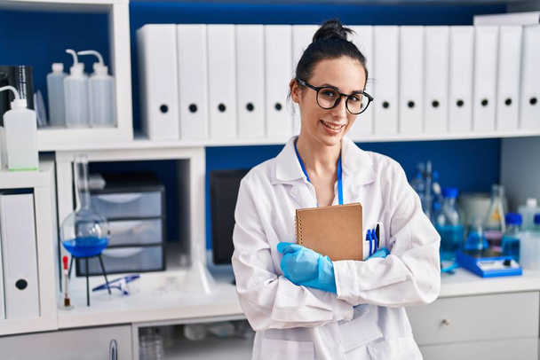 Junge kaukasische Wissenschaftlerin lächelt zuversichtlich und hält Notizbuch im Labor - Foto, Bild