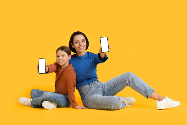 Wesoła matka i mała córeczka demonstrujące smartfony z białym ekranem przed kamerą, szczęśliwa mama i dziecko zaleca nową aplikację mobilną siedząc na żółtym tle, makieta - Zdjęcie, obraz
