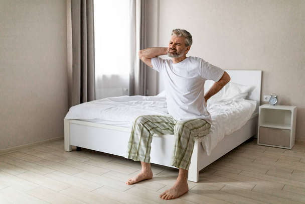 Tired mature man suffering from back pain after sleeping, waking up in morning, sitting on bed and touching his lower back and neck, experiencing body aches, wearing pajamas, copy space, full length - Foto, imagen