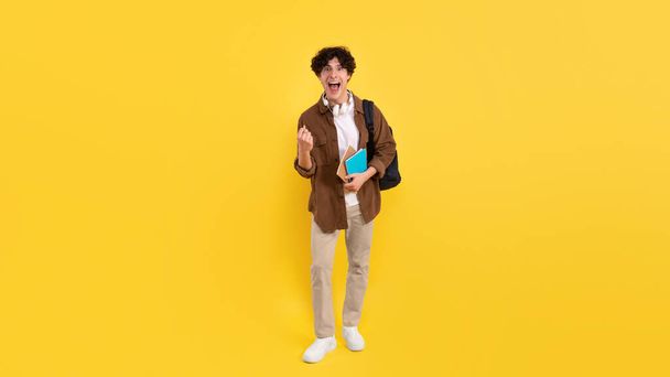 Emotional Student Guy Shouting Gesturing Yes Celebrating Educational Luck Entering University Posing With Backpack And Notebooks Over Yellow Studio Background, Looking At Camera. Panorama - Photo, Image