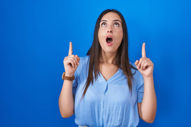 Young brunette woman standing over blue background amazed and surprised looking up and pointing with fingers and raised arms.  - Photo, Image