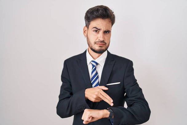 Junger hispanischer Mann mit Tätowierungen, der eilig Geschäftsanzug und Krawatte trägt und auf die Zeit verweist, ungeduldig, aufgebracht und wütend auf die Fristverlängerung  - Foto, Bild