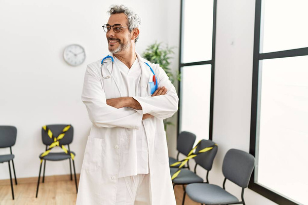 Homme moyen aux cheveux gris médecin souriant confiant debout avec les bras croisés geste dans la salle d'attente de la clinique - Photo, image
