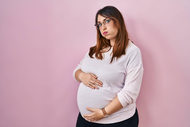 Zwangere vrouw staat over roze achtergrond puffende wangen met grappig gezicht. mond opgeblazen met lucht, gekke uitdrukking.  - Foto, afbeelding