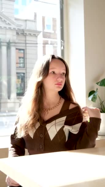 Mädchen auf dem Hintergrund eines Fensters in einer braunen Jacke in einem Café junge Frau im Alter von 13-16 Jahren schaut in den Rahmen der Haar- und Hautpflege - Filmmaterial, Video