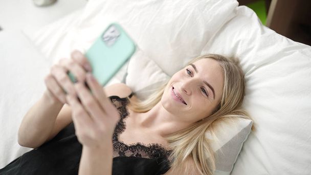 Giovane donna bionda utilizzando smartphone sul letto in camera da letto - Foto, immagini