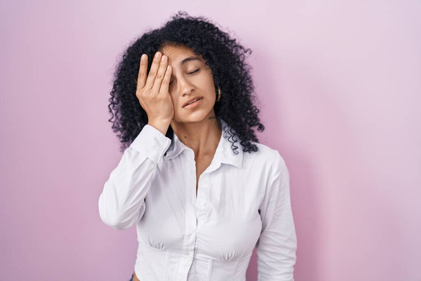 Hispanic woman with curly hair standing over pink background yawning tired covering half face, eye and mouth with hand. face hurts in pain.  - Fotografie, Obrázek