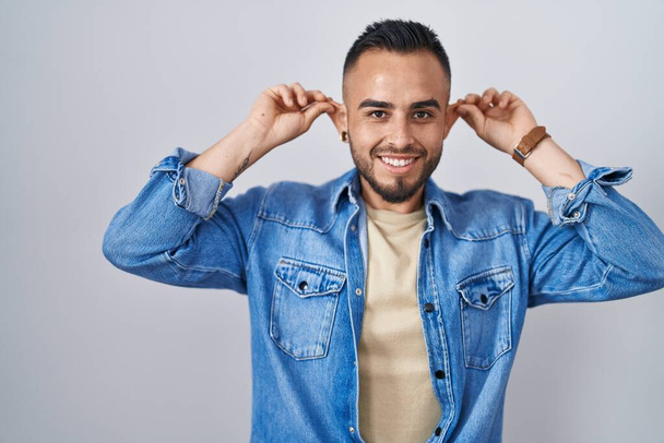 Young hispanic man standing over isolated background smiling pulling ears with fingers, funny gesture. audition problem  - Fotoğraf, Görsel