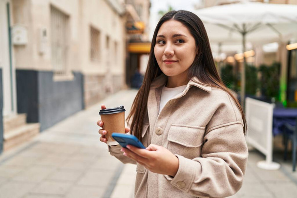 Young hispanic woman using smartphone drinking coffee at street - Valokuva, kuva