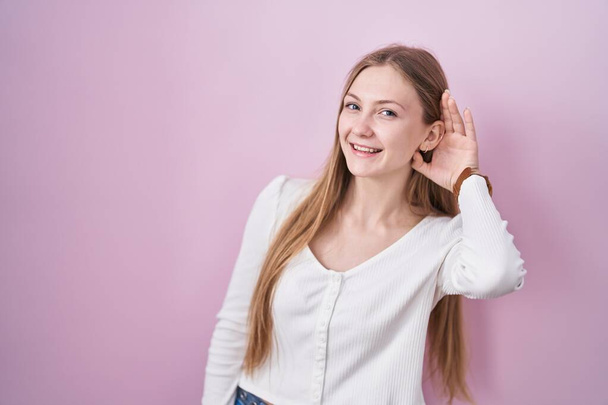 Young caucasian woman standing over pink background smiling with hand over ear listening an hearing to rumor or gossip. deafness concept.  - Photo, image