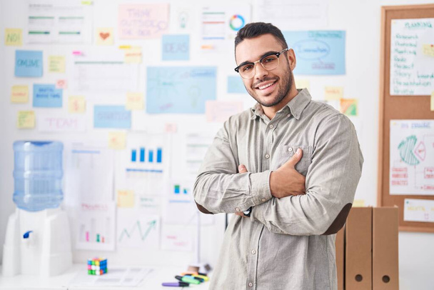 Νεαρός ισπανόφωνος επιχειρηματίας χαμογελά σίγουρος στέκεται με τα χέρια σταυρωμένα χειρονομία στο γραφείο - Φωτογραφία, εικόνα