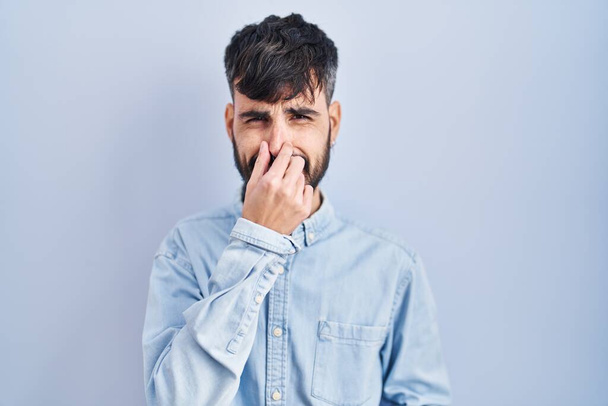 Jeune homme hispanique avec la barbe debout sur fond bleu sentant quelque chose de puant et dégoûtant, odeur intolérable, retenant souffle avec les doigts sur le nez. mauvaise odeur  - Photo, image