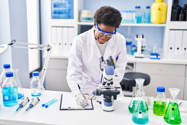 Αφροαμερικανή γυναίκα φορώντας στολή επιστήμονα χρησιμοποιώντας μικροσκόπιο γράψει στο πρόχειρο στο εργαστήριο - Φωτογραφία, εικόνα