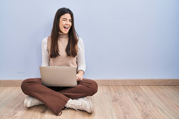 Young brunette woman working using computer laptop sitting on the floor winking looking at the camera with sexy expression, cheerful and happy face.  - Foto, afbeelding