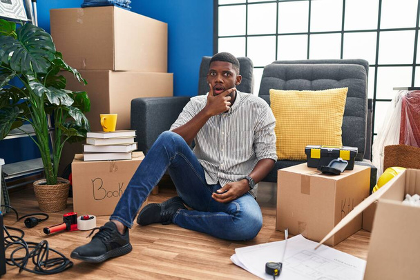 Uomo afroamericano seduto sul pavimento in una nuova casa cercando affascinato con incredulità, sorpresa e espressione stupita con le mani sul mento  - Foto, immagini