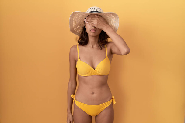 Jonge Spaanse vrouw met bikini en zomerhoed die de ogen met de hand bedekt en er ernstig en verdrietig uitziet. gezichtsloosheid, verstoppertje en afwijzing  - Foto, afbeelding