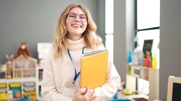 Молода блондинка вчителька дошкільного віку посміхається впевнено тримає книги в дитячому садку
 - Фото, зображення