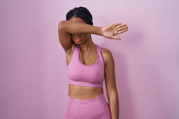 Африканская американка с косичками в спортивной одежде на розовом фоне закрывает глаза рукой, выглядит серьезным и грустным. беспросветная, скрывающая и отвергающая концепция  - Фото, изображение