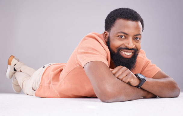 Portrait, happy and a black man lying on the floor isolated on a grey studio background. Smile, relax and a calm African guy relaxing, smiling and looking confident on the ground of a backdrop. - Photo, image