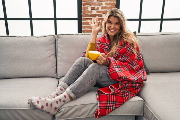 Young woman sitting on the sofa drinking a coffee at home smiling positive doing ok sign with hand and fingers. successful expression.  - Foto, Imagen
