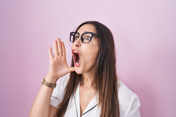 Jonge brunette vrouw met een bril over roze achtergrond schreeuwend en schreeuwend met hand op mond. communicatieconcept.  - Foto, afbeelding