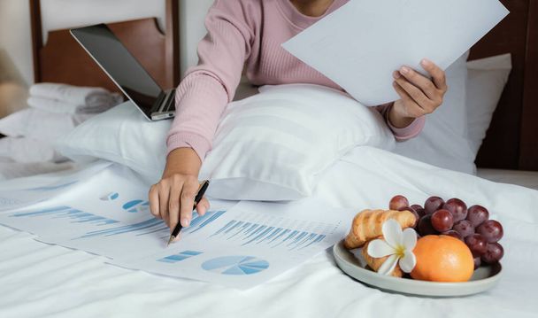young woman hand holding pen used laptop video conferences and eating fruits on the bed, healthy food and work from home concept. - 写真・画像